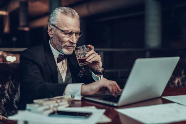 Businessman Using Laptop. Old Businessman . Man is Wearing in Black Suit. Experienced Entrepreneur. Strict Man. looking at Laptop. stack of money on table. holding a glass of cognac.