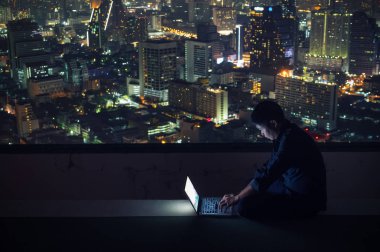 Asya işadamı oturmak ve dizüstü bilgisayarlar ile arka planında bulanık gökdelenler, gece kullanmak iş başarı ve teknoloji kavramları.