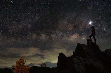Dağcı veya backpacker silüeti. Elinde, o başını yüksek ışık düzenlenen. Kayalık dağ tepe ve Samanyolu Gökadası, başarı, kazanan, lider kavramı üzerinde duruyor. Yüksek ISO gürültü ile.