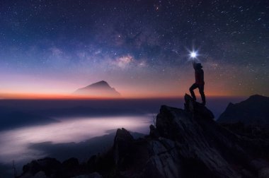 Dağcı veya backpacker silüeti. Elinde, o başını yüksek ışık düzenlenen. Kayalık dağ tepe ve Samanyolu Gökadası, başarı, kazanan, lider kavramı üzerinde duruyor. Yüksek ISO gürültü ile.
