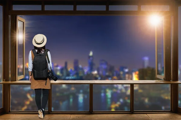 Asian female tourist, She wears casual style Japanese.with simple black backpack and camera with lens behind looking cityscape view, Standing behind cafe counter, looking out bangkok city view.