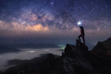 Dağcı veya backpacker silüeti. Elinde, o başını yüksek ışık düzenlenen. Kayalık dağ tepe ve Samanyolu Gökadası, başarı, kazanan, lider kavramı üzerinde duruyor. Yüksek ISO gürültü ile.