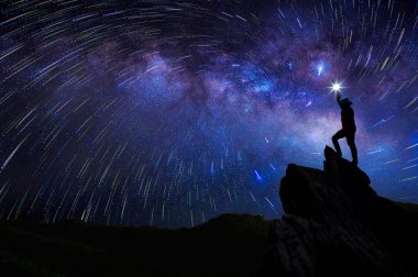 Dağcı veya backpacker silüeti. Elinde, o başını yüksek ışık düzenlenen. Kayalık dağ tepe ve Samanyolu Gökadası, başarı, kazanan, lider kavramı üzerinde duruyor. Yüksek ISO gürültü ile.