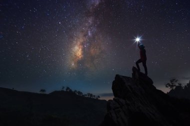 Dağcı veya backpacker silüeti. Elinde, o başını yüksek ışık düzenlenen. Kayalık dağ tepe ve Samanyolu Gökadası, başarı, kazanan, lider kavramı üzerinde duruyor. Yüksek ISO gürültü ile.