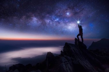 Dağcı veya backpacker silüeti. Elinde, o başını yüksek ışık düzenlenen. Kayalık dağ tepe ve Samanyolu Gökadası, başarı, kazanan, lider kavramı üzerinde duruyor. Yüksek ISO gürültü ile.