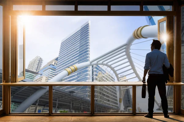 Concept vision, Young businessman wearing comfortable casual suit jacket standing in working room near the glass window and looking through the modern buildings in the skyline.