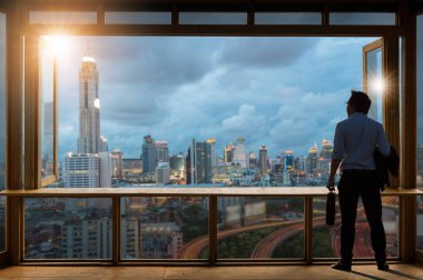 Kavram görme, cam pencere yakınındaki çalışma odasında duran ve manzarası modern binalarda bakarak rahat rahat elbise giyen genç işadamı.