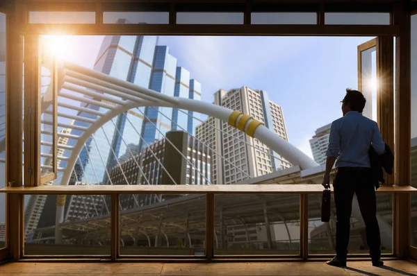Concept vision, Young businessman wearing comfortable casual suit jacket standing in working room near the glass window and looking through the modern buildings in the skyline.
