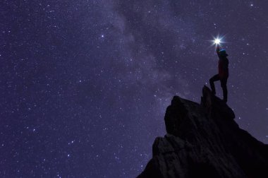 Silhouette of climber or backpacker, he held the light up high above his head. Standing on rocky mountain peak against Milky way galaxy, stars and space dust in the universe, success, leader concept.
