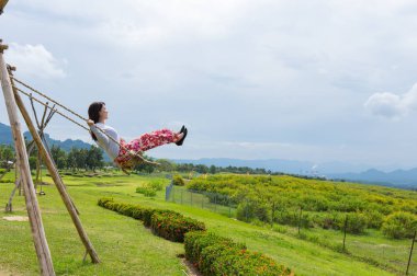 Woman wearing red dress sitting on swing on clear day in Tung Bua Tong Mexican sunflower field in Mae Moh Coal Mine, Lampang Province, Thailand. clipart