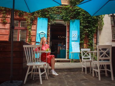 Elbiseli genç bir kadın, eski bir Avrupa şehrinin caddesindeki bir yazlık kafede bir buket çiçekle oturuyor.