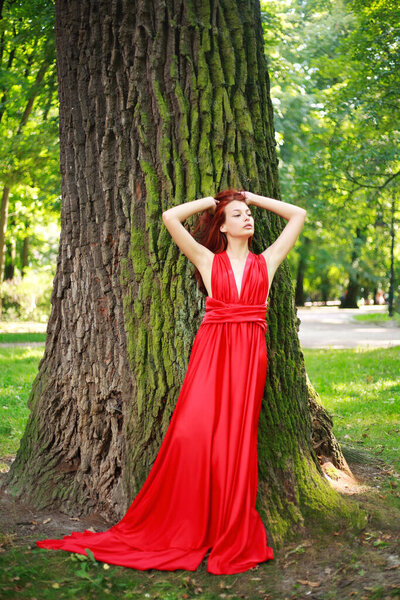 Attractive young girl, lady in a long red dress in a summer forest park, fashionable woman outdoors in nature