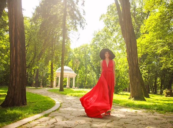 Uzun kırmızı elbiseli, şık bir kadın yaz parkında yürüyor.
