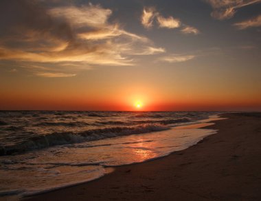 Gökyüzünde güzel bir gün batımı, deniz kıyısında kumlu bir sahil.