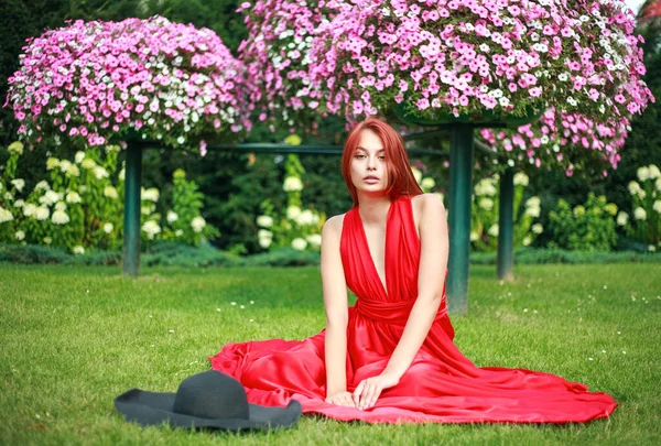 芝生の上の花畑で黒い帽子をかぶった長い赤いドレスの若い魅力的な女の子 — ストック写真