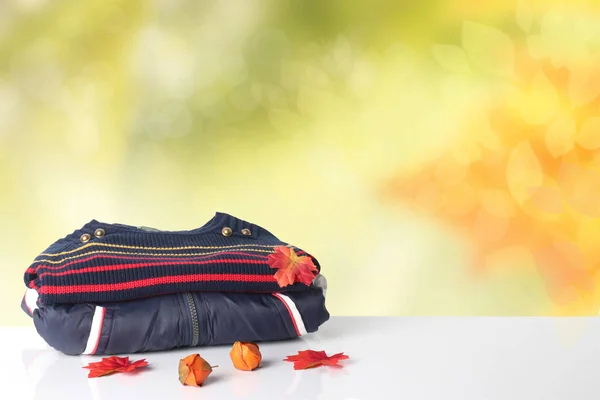 Stack colorful children autumn clothes. Pile of colorful warm children clothes on white table in front of abstract blurred bright yellow autumn background. Space for your display product montage. Autumn fashion.