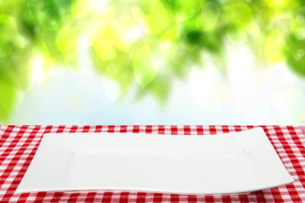 Empty plate background. Empty white plate on table with red napkin or tablecloth over abstract natural summer background. Template for your food and product display montage. Concept food. Copy space.