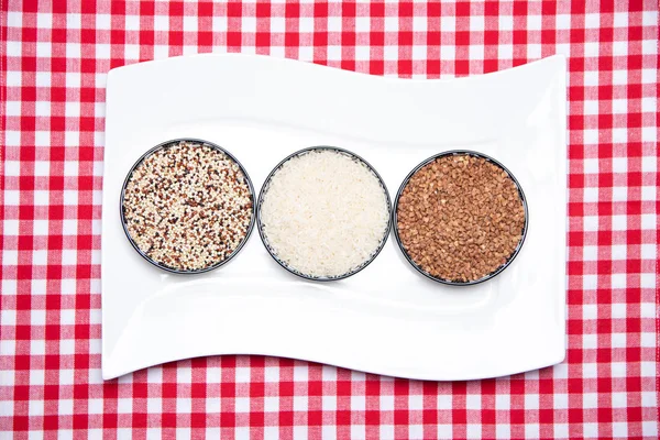 Vegetable food. A decorative white plate with three bowls with buckwheat, rice and quinoa on a red checkered tablecloth. Healthy eating. Top view.