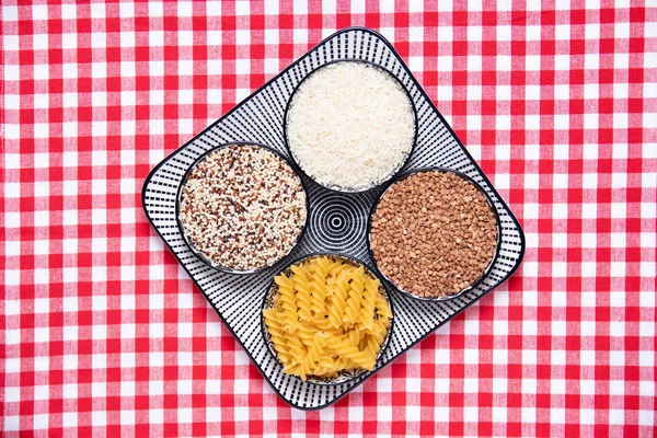 Vegetable food. A decorative plate with four bowls with buckwheat, noodles, rice and quinoa on a red checkered tablecloth. Healthy eating. Top view with space for design.