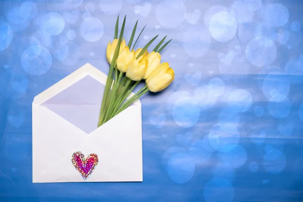 Happy Valentine or Mothers Day background. Beautiful bouquet of yellow tulips in an open envelope with a hearts symbol lying on a abstract blue background. Space for your design on the right side.