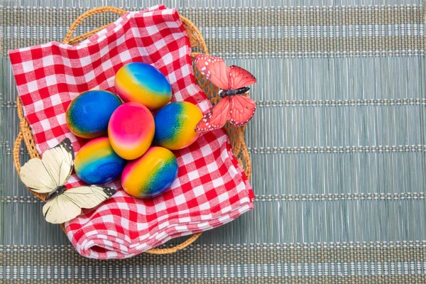 Feliz Pascua de fondo. Vista superior de coloridos huevos de Pascua en una canasta en una servilleta a cuadros roja sobre una esterilla de bambú verde. Espacio para su diseño . — Foto de Stock