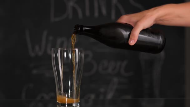 Pouring cool beer from bottle into glass on dark wall background. — Stock Video