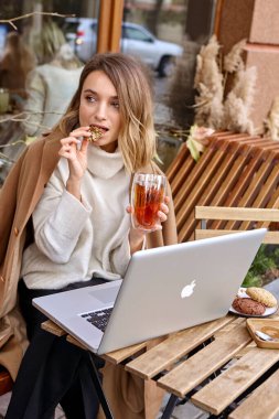 Kherson, Kherson Bölgesi, Ukrayna - 24 Ekim 2019: Bir kadın elinde dizüstü bilgisayarla bir fincan kahve ve bir telefon eşliğinde tatlı bir tatlıyla masada oturuyor.