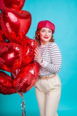 Stüdyoda mavi arka planda kalp şeklinde şişme kırmızı balonları olan bir kadın.