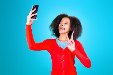 Stüdyoda mavi arka planda tıbbi maskeli ve telefonu olan esmer tenli bir kadın.