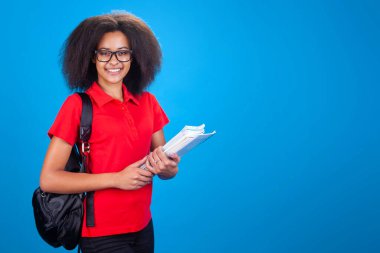 Elinde çanta ve belgelerle esmer tenli bir bayan öğrenci stüdyoda mavi bir altyapıya sahip.