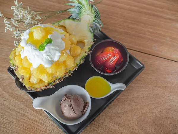 Ice Cream Snow Flake. Sweet Dessert Korean Style call Bingsu with Pineapple Flavor Ice Ball in A Bowl Made by A Half of Fresh Pineapple, Jelly in Black Cup, Chocolate Ice Cream in White Cup, Pineapple Flavor Sauce in White Ceramic Spoon on Wooden Tab