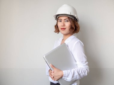 Güzel Asyalı kadın mühendislik kıvırcık kısa saç beyaz kask ve gümüş laptop beyaz duvar arka planda tutan beyaz gömlek giyiyor. Çalışan kadın kavramı.