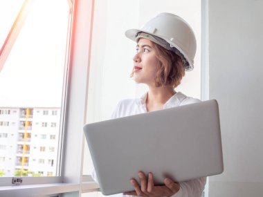 Beyaz kask ve İnşaat Sanayi ile cam pencere yakınındaki gümüş dizüstü bilgisayar ile çalışan beyaz gömlek giymiş güzel Asyalı kadın mühendis kıvırcık kısa saçlı. Çalışan kadın kavramı.