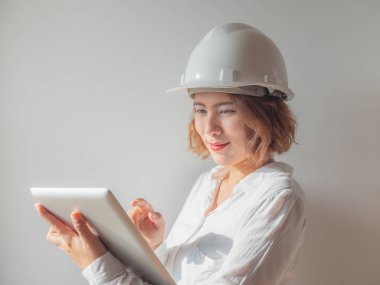 Beyaz kask ve gümüş tablet ile çalışma ve gülümseyen beyaz gömlek giymiş güzel Asyalı kadın mühendis kıvırcık kısa saçlı. Çalışan kadın kavramı.