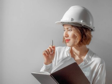 Güzel Asyalı kadın mühendislik kıvırcık kısa saç beyaz kask ve defter ve gülümseyen gümüş kalem tutan beyaz gömlek giyiyor. Çalışan kadın kavramı.
