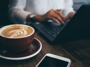 El bilgisayar fare, kafede ahşap masa üzerinde kahve ve cep telefonu ile çalışma konusunda yoğunlaşmış olan siyah dizüstü bilgisayarda yazarak. Çalışan kadın kavramı.