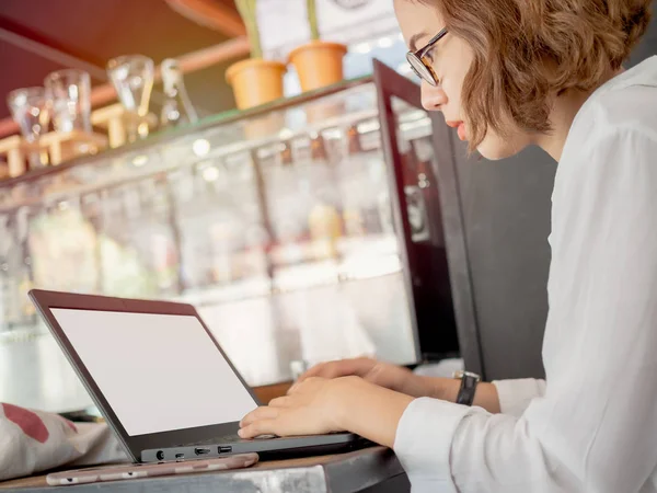 Güzel Asyalı kadın gözlük ve beyaz gömlek giymiş siyah dizüstü bilgisayar ve cep telefonu kafede ahşap masa üzerinde yazarak çalışma, konsantre. Çalışan kadın kavramı.