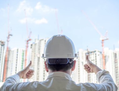 Mühendis adam geri görüntülemek zor Emanet kask ve beyaz gömlek gösteren başparmak yukarıya için başarı ile her ikisi el giyen beyaz güneş gününde arka bina inşaat.