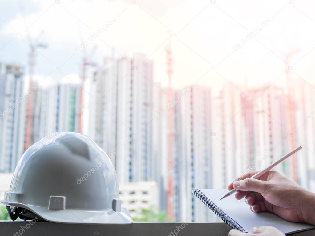 White hard safety helmet for civil engineer, protect and safety hat for engineering worker and hand writing notebook by pencil on construction building site background.