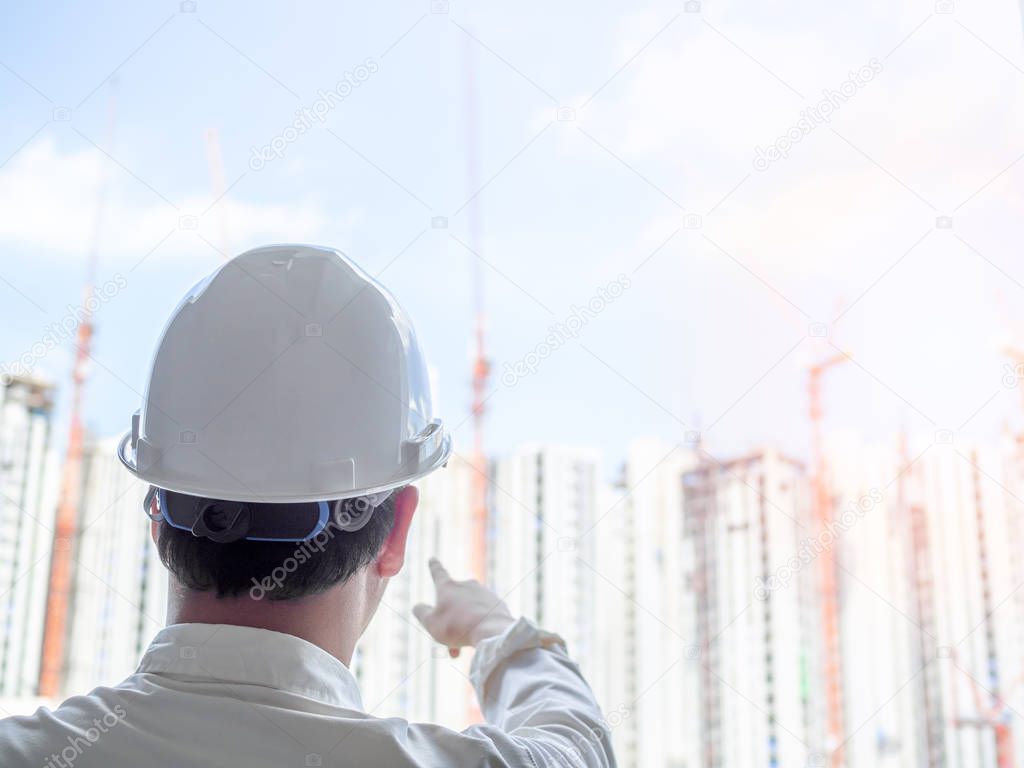 Engineer man back view wearing white hard safety helmet and white shirt pointing his right hand on construction building background on sunshine day.