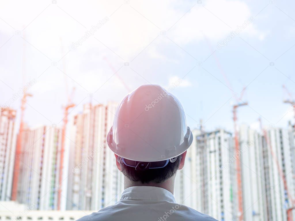 Engineer man back view wearing white hard safety helmet and white shirt looking to construction building background on sunshine day.