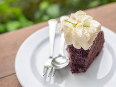 Tepesi ve kırbaçlanan krem beyaz seramik plaka üstüne sillver kaşık ve çatalı yeşil doğa arka plan üzerinde ahşap masa güzel vintage çiçekler ile vahşi organik çikolatalı bal pasta bir parça.