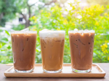Three glasses of iced coffee on wooden table on green garden background in cafe. clipart