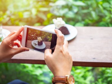 Çikolatalı Pasta Cafe yeşil bahçe zemin üzerine bir akıllı telefon kameradan ahşap masa üzerinde beyaz plaka üzerinde çekim eller.