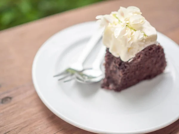 A piece of wild organic chocolate honey cake with beautiful vintage flowers topping and whipping cream on top in white ceramic plate with sillver spoon and fork on wooden table on green nature background.