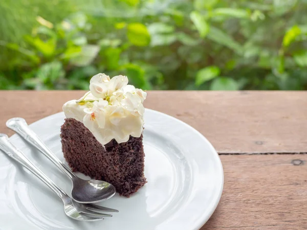 A piece of wild organic chocolate honey cake with beautiful vintage flowers topping and whipping cream on top in white ceramic plate with sillver spoon and fork on wooden table on green garden background.