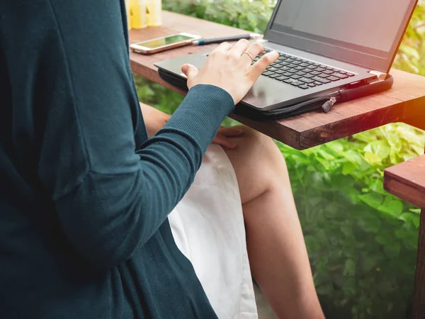 Woman wearing blue shirt and white skirt typing laptop computer on wooden counter bar and sitting cross legs near the window with garden view in the cafe.