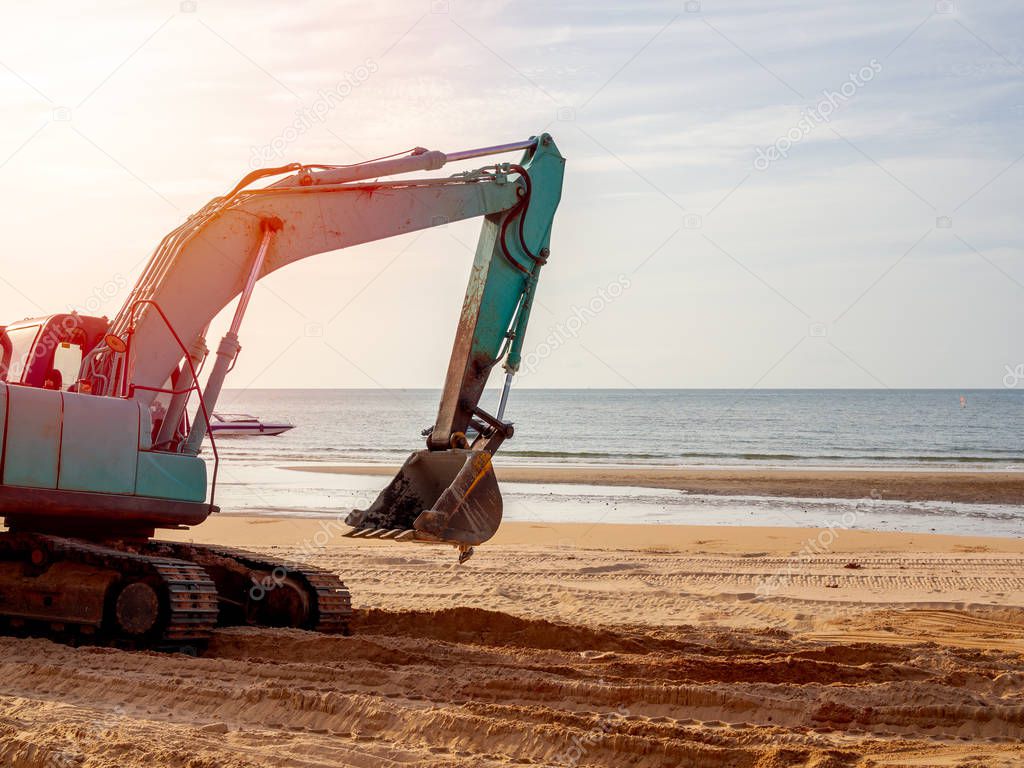 Blue or green Excavator working on the beach with sea view on sunset. Excavator Tire track on sand with sunlight in the evening on seascape background.