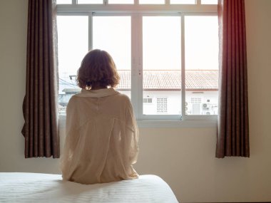 Kadın kısa saç beyaz yatakta otel odasında oturuyor. Yalnız kadın beyaz otel odasında tek başına pencere bakarak.