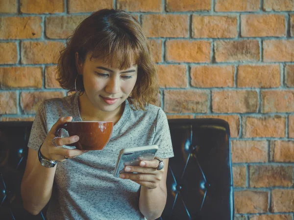 Beautiful asian woman short hair smiling and holding a cup of coffee and looking mobile phone and sitting on black vintage sofa on brick wall background with copy space.